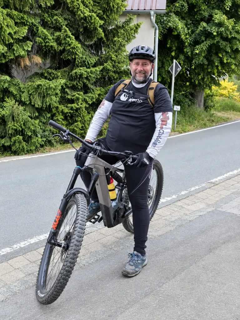 Shredwoodi lächelt mit Helm und Rucksack in die Kamera und hält sein E-Mountainbike am Straßenrand.